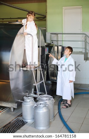 Smiling girl and boy in white coats stand near large tank of milk at factory.