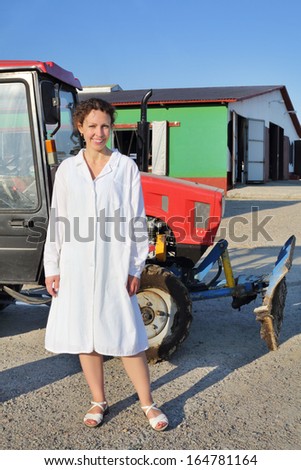 Happy woman in white robe stands near red tractor in big farm at sunny day.