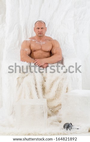A strong man with big muscles with snow on body sitting in snow chair with fur in studio