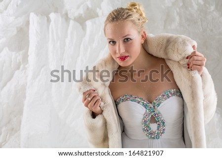 Beautiful girl with snow on face in white dress and fur coat in studio with snow wall