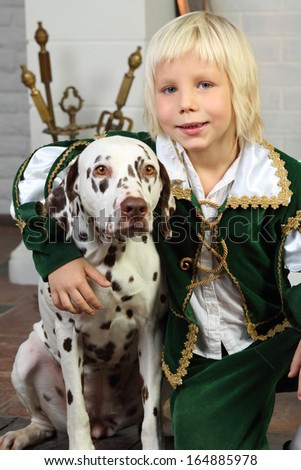 Happy blonde little boy in medieval costume sits with dalmatian near fireplace.