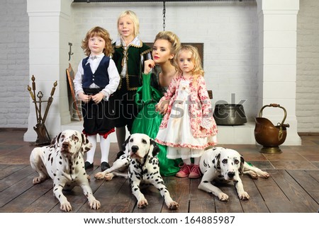 Beautiful woman in green medieval costume with three children and three dalmatians near fireplace.
