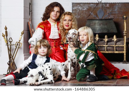 Happy father, mother and two sons in medieval costumes sit with two dalmatians near fireplace.