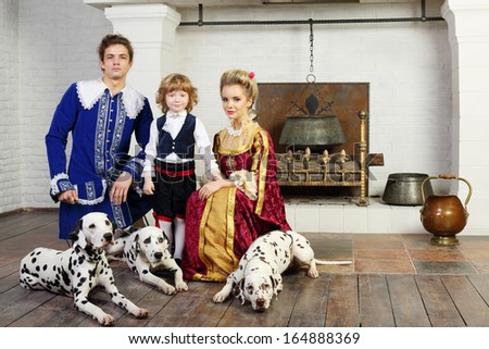 Young father, mother and little son in medieval costume stand near fireplace with three dalmatians on leashes.