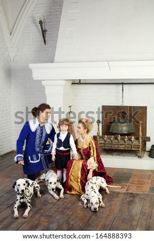Young father, mother in medieval costumes look at little son near fireplace with three dalmatians on leashes.