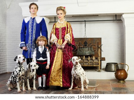 Father, mother and happy little son in medieval costume stand near fireplace with three dalmatians on leashes.
