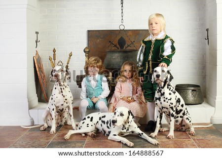Two boys and girl in medieval costumes sit near fireplace with hanging pot with dalmatians.