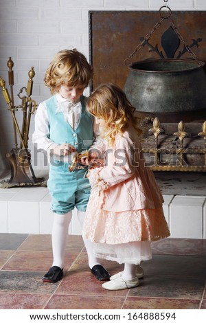 Little boy and girl in medieval costumes look at small casket near fireplace with boiler.