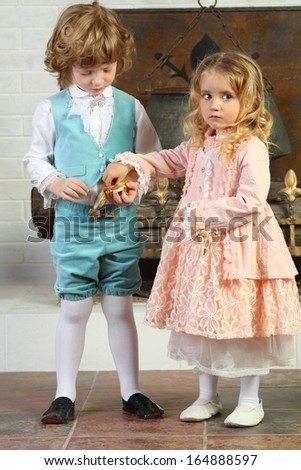 Little boy and girl in medieval costumes play with small casket near fireplace with boiler.
