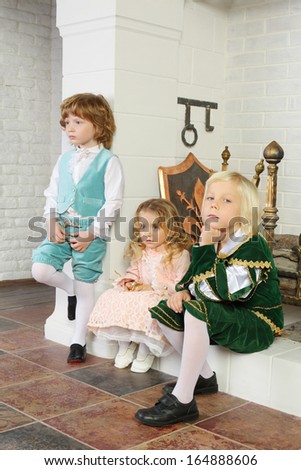 Two boys and girl in medieval costumes sit near fireplace with logs. Focus on left boy and girl.