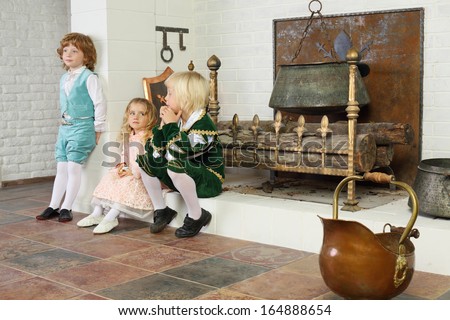 Two boys and girl in medieval costumes sit near fireplace with logs and hanging pot.