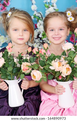 Two little girls in dresses hold bouquets of roses in pitchers and smile in room with flowers. Focus on right girl.