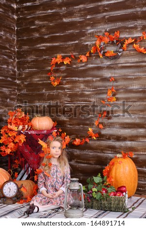 Little girl sits on floor with old lamp and basket of fruits in room with pumpkins and autumn interior.