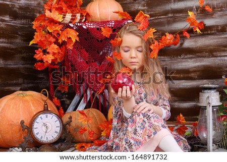 Pretty little girl sits on floor and looks at red apple in room with pumpkins and autumn interior.