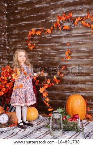 Little girl holds garaland of orange leaves in room with pumpkins and autumn interior.
