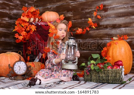 Pretty little girl sits on floor and blows out old lamp in room with pumpkins and autumn interior.