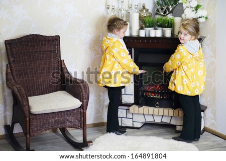 Two little girls in same clothes stands near fireplace and wicker rocking chair in cosy room. Focus on right girl.