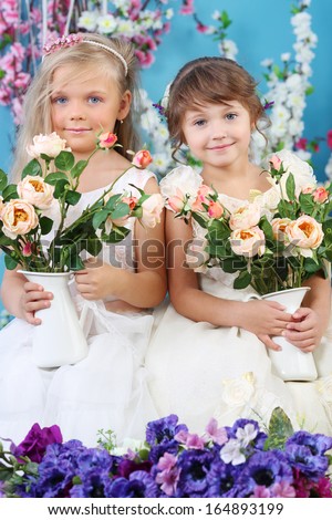 Two pretty little girls in white dresses hold bouquets of roses and smile in room with flowers.