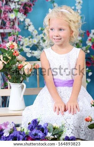 Blonde little girl in white dress sits on bench near bouquet of roses and looks away in room with flowers.