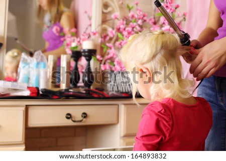 Back of pretty blonde little girl and hands of hairdresser with curling iron.