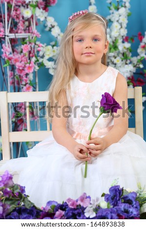 Pretty little girl in white dress sits and holds violet rose in room with flowers.