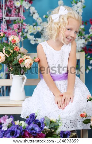 Blonde little girl in white dress sits on bench near bouquet of roses and nicely smiles in room with flowers.