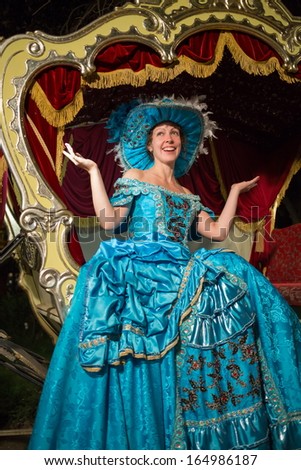 The smiling girl in blue old-fashioned dress and hat sits in a carriage with raised hands