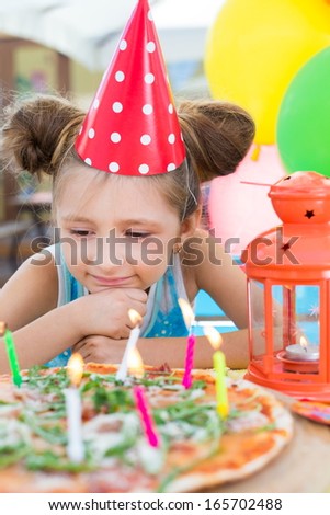 Girl in festive cap with candles in pizza on table in cafe