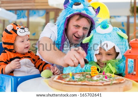 Father with daughter in monster costumes and baby boy in tiger costume celebrate the birthday in a cafe, father ignites candles.