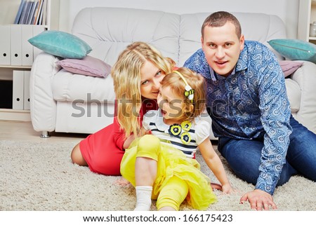 Family of three sits on carpet in light room, daughter ahead, parents behind, mother whispers something at daughter ear