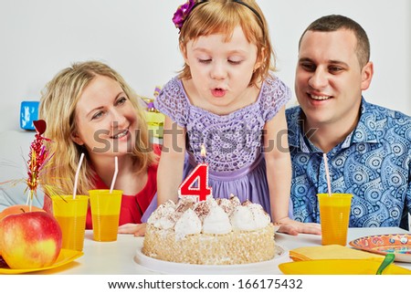 Family of three sits at birthday table, daughter blows out candle on cake and parents look at her