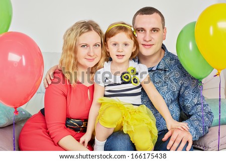 Family of three sits on sofa in light room decorated with birthday air balloons, daughter sits on father knee