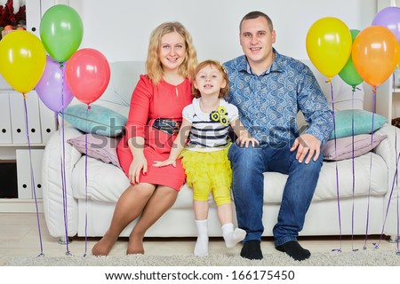 Family of three sits on sofa in light room decorated with birthday air balloons