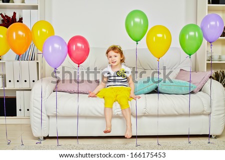 Little girl sits on big sofa in room with carpet on floor and birthday air balloons