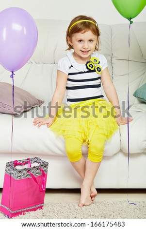 Little barefooted girl sits on big sofa in room with carpet on floor, paper gift bag and birthday air balloons