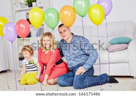 Family of three sits on carpet in light room behind row of birthday balloons