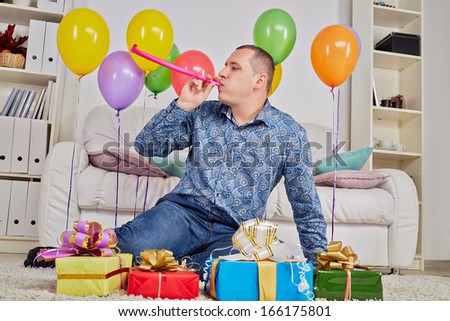 Man sits on carpet among gift boxes and birthday air balloons and blows into party-blower