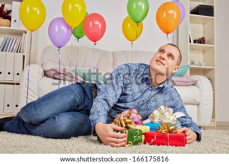 Man lies on carpet among gift boxes and birthday air balloons and looks dreamily at ceiling