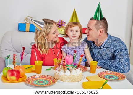 Mother, father and little daughter sit together on  light soft sofa at birthday table