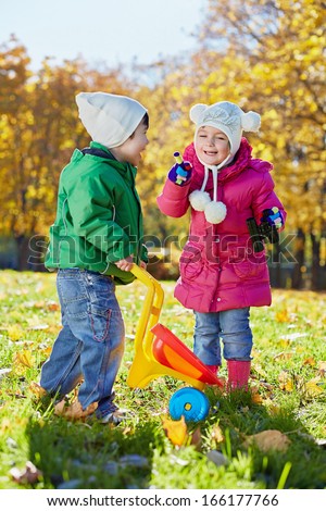 Little boy with plastic wheelbarrow and little girl with binocular cheerfully talk at sunny dlade in autumn park
