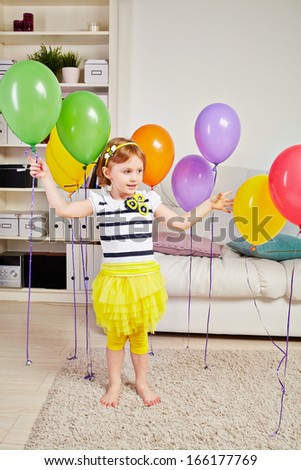 Little girl stands in room on light carpet on floor and touches birthday air balloons