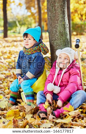 Little children sit leaning at tree trunk in autumn park, boy sits on rucksack, girl sits on push-scooter