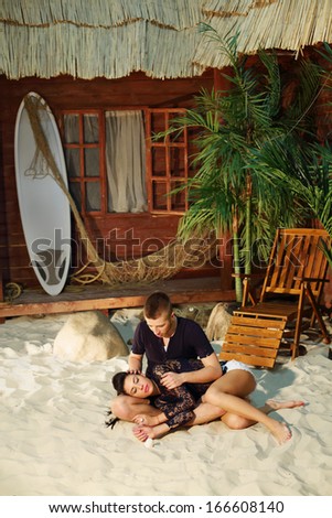 Man sits on sand and woman lies on his lap next to beach house.