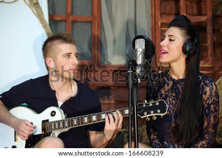 Man plays guitar and woman sings next to beach house. Man looks at woman. Focus on woman.