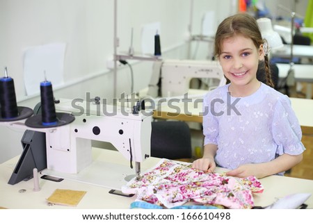 Smiling girl stands near table with sewing machine in classroom with many machines.
