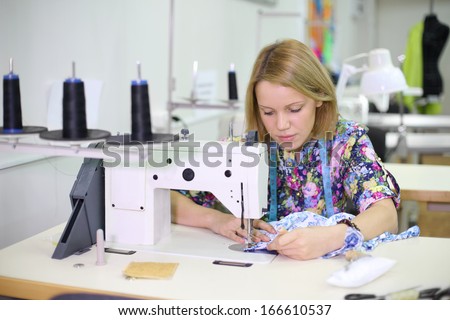 Pretty female tailor sits at table with sewing machine and sews fabric.