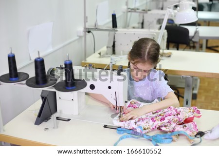 Student girl sews small dress at white sewing machine in classroom with many machines.