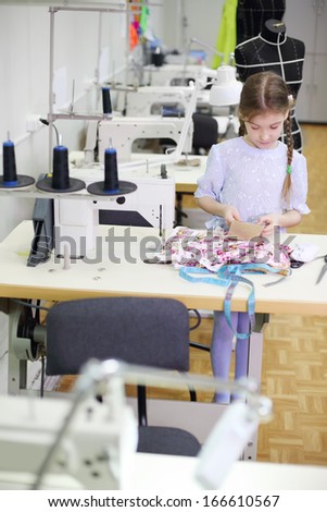 Girl stands near table with sewing machine and holds cloth in classroom with many machines.