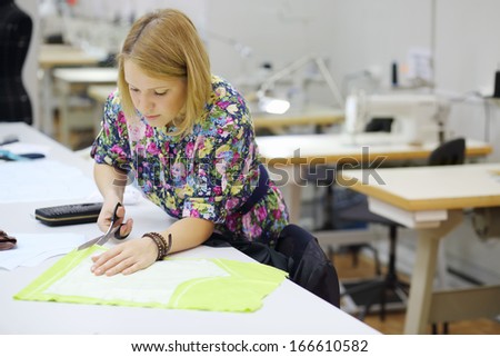 Pretty female tailor stands near table and cuts tailoring detail at light green cloth.