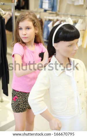 Little pretty girl in shorts touches hair of dummy and looks away in children store.